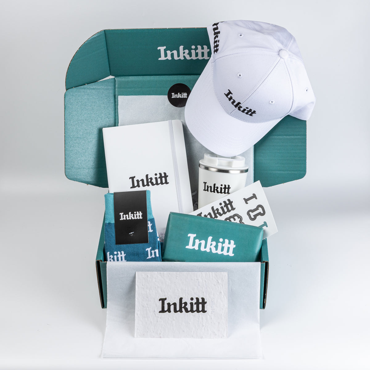 An Inkitt gift box set featuring a white baseball cap, a white notebook, a white mug, a teal t-shirt, and matching teal and white accessories, all neatly arranged inside and in front of an open teal box on a light gray background.